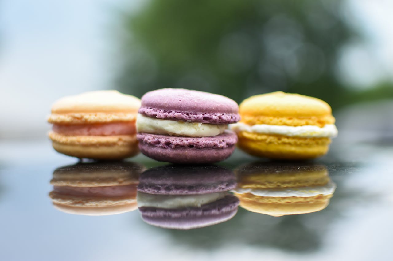 Vibrant macarons with cream filling against blurred greenery, showcasing reflections on a shiny surface.